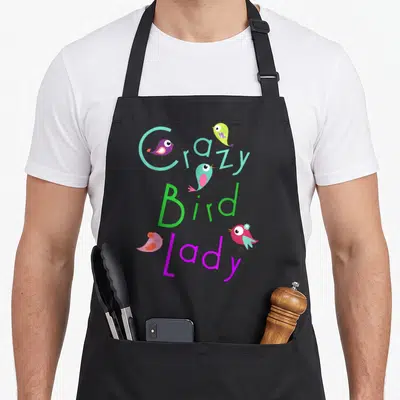 Funny pet bird apron - Crazy Bird Lady text with four colorful cartoon birds and a blue flower accent — shown on a black cotton canvas apron against a white background