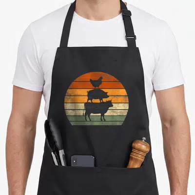 Cow pig farm aprons - Stacked chicken pig cow silhouettes on orange yellow green grey striped retro sunset circle — shown on a black cotton canvas apron against a white background