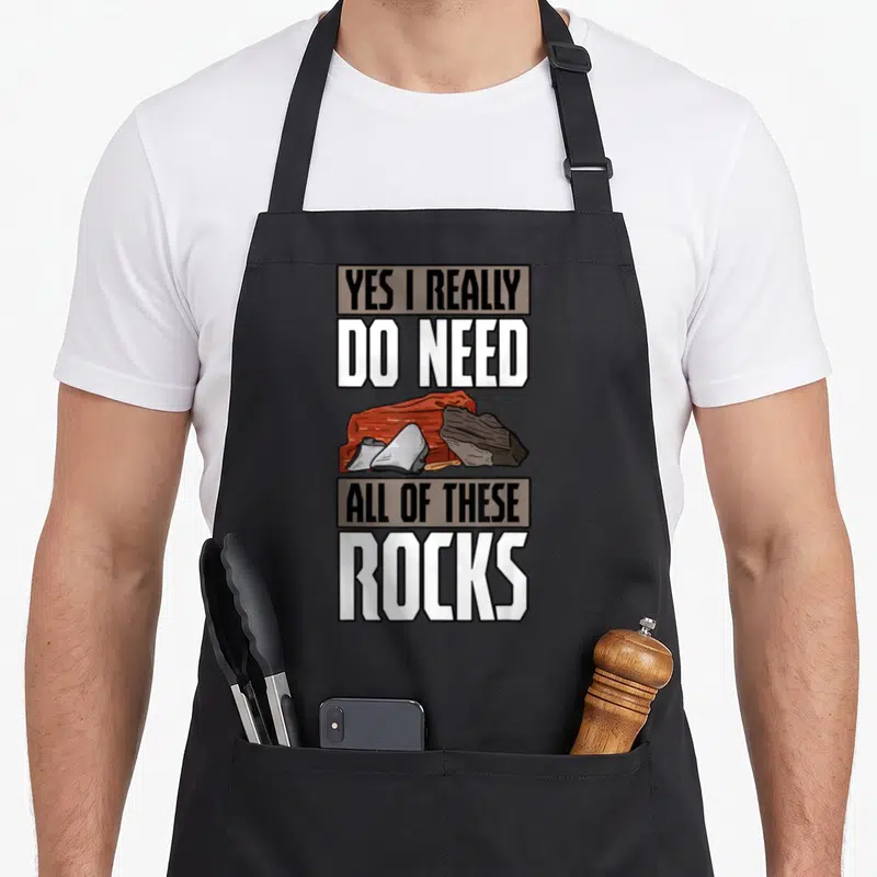 Earth geologist apron - Cartoon rocks in brown grey and white surrounded by text YES I REALLY DO NEED ALL OF THESE ROCKS — shown on a black cotton canvas apron against a white background