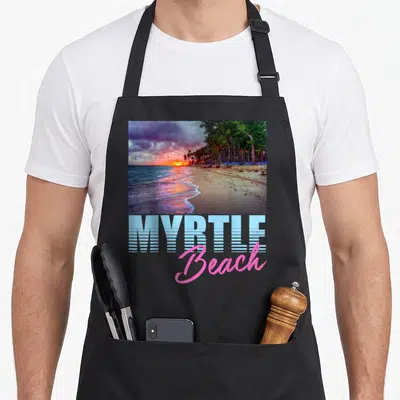 Ocean pride apron - Rectangular sunset beach photo above blue block and pink script MYRTLE Beach text — shown on a black cotton canvas apron against a white background