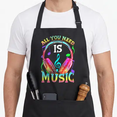Headphone producer apron - Cartoon headphones with All You Need Is Music text in cyan magenta yellow and white — shown on a black cotton canvas apron against a white background