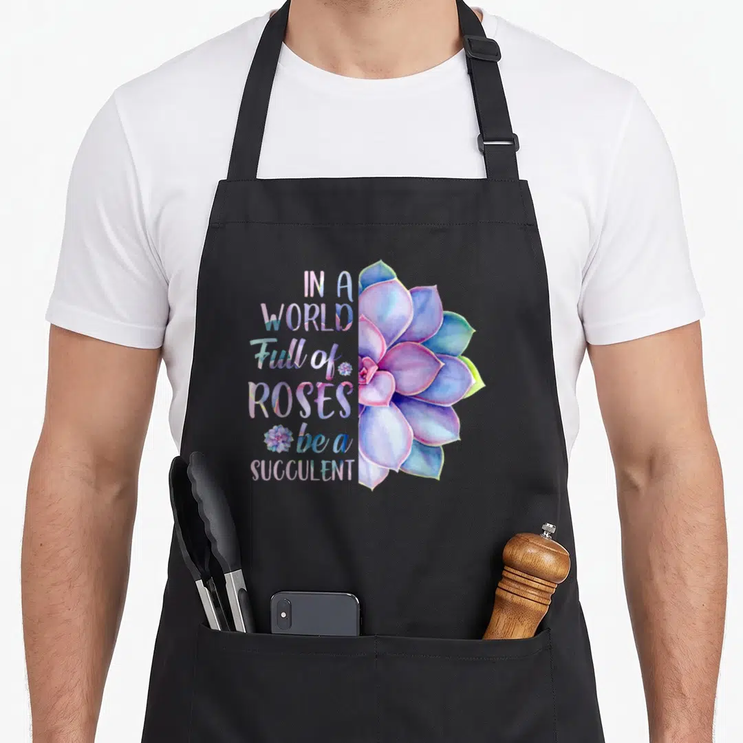 Succulent rose apron - Watercolor succulent in purple and blue next to stacked text reading IN A WORLD Full of ROSES be — shown on a black cotton canvas apron against a white background