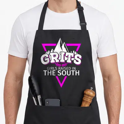 South grit southern aprons - Pink triangle frame enclosing white mountains and GRITS GIRLS RAISED IN THE SOUTH text — shown on a black cotton canvas apron against a white background