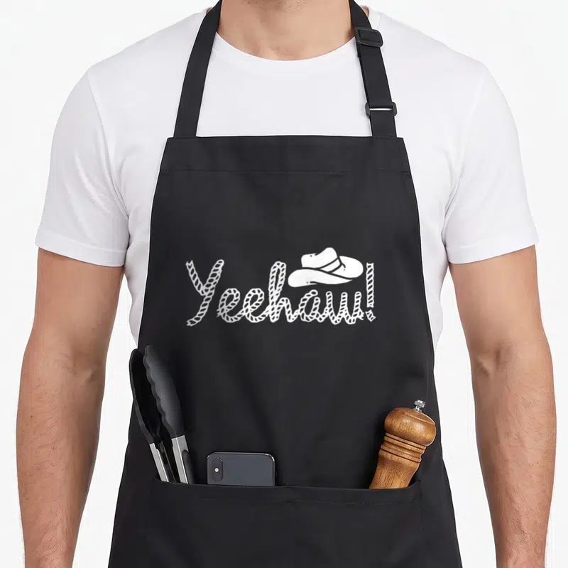 Square cook's country aprons - White Yeehaw rope text with cowboy hat on black bib — shown on a black cotton canvas apron against a white background