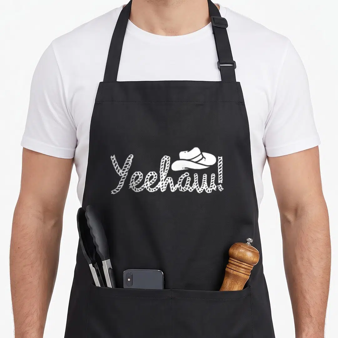 Square cook's country aprons - White Yeehaw rope text with cowboy hat on black bib — shown on a black cotton canvas apron against a white background