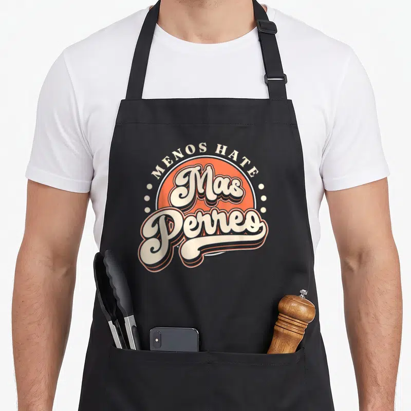 Dance modern apron - Retro orange circle with MENOS HATE Mas Perreo text in off-white and black script — shown on a black cotton canvas apron against a white background