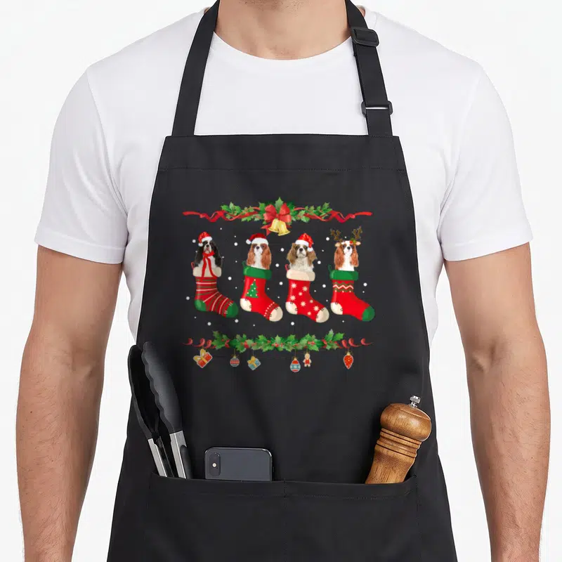 Cavalier womens christmas aprons - Four cartoon spaniels in red and green stockings with holly garland and holiday — shown on a black cotton canvas apron against a white background