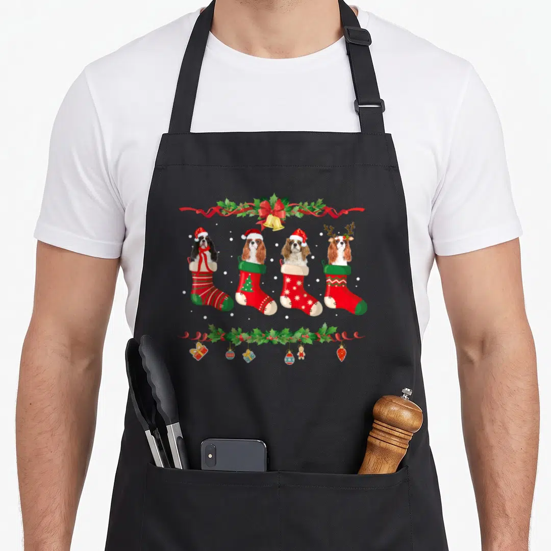 Cavalier womens christmas aprons - Four cartoon spaniels in red and green stockings with holly garland and holiday — shown on a black cotton canvas apron against a white background