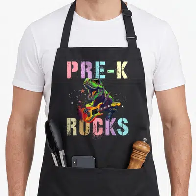 Pre rock teacher apron - Green T-Rex playing orange guitar with PRE-K ROCKS text in pastel colors — shown on a black cotton canvas apron against a white background