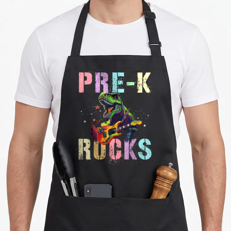 Pre rock teacher apron - Green T-Rex playing orange guitar with PRE-K ROCKS text in pastel colors — shown on a black cotton canvas apron against a white background
