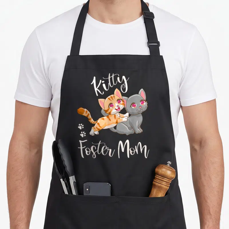 Foster kitty cat apron - Two cartoon kittens orange and grey with arched text and paw prints — shown on a black cotton canvas apron against a white background
