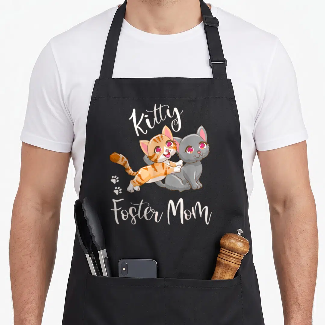 Foster kitty cat apron - Two cartoon kittens orange and grey with arched text and paw prints — shown on a black cotton canvas apron against a white background
