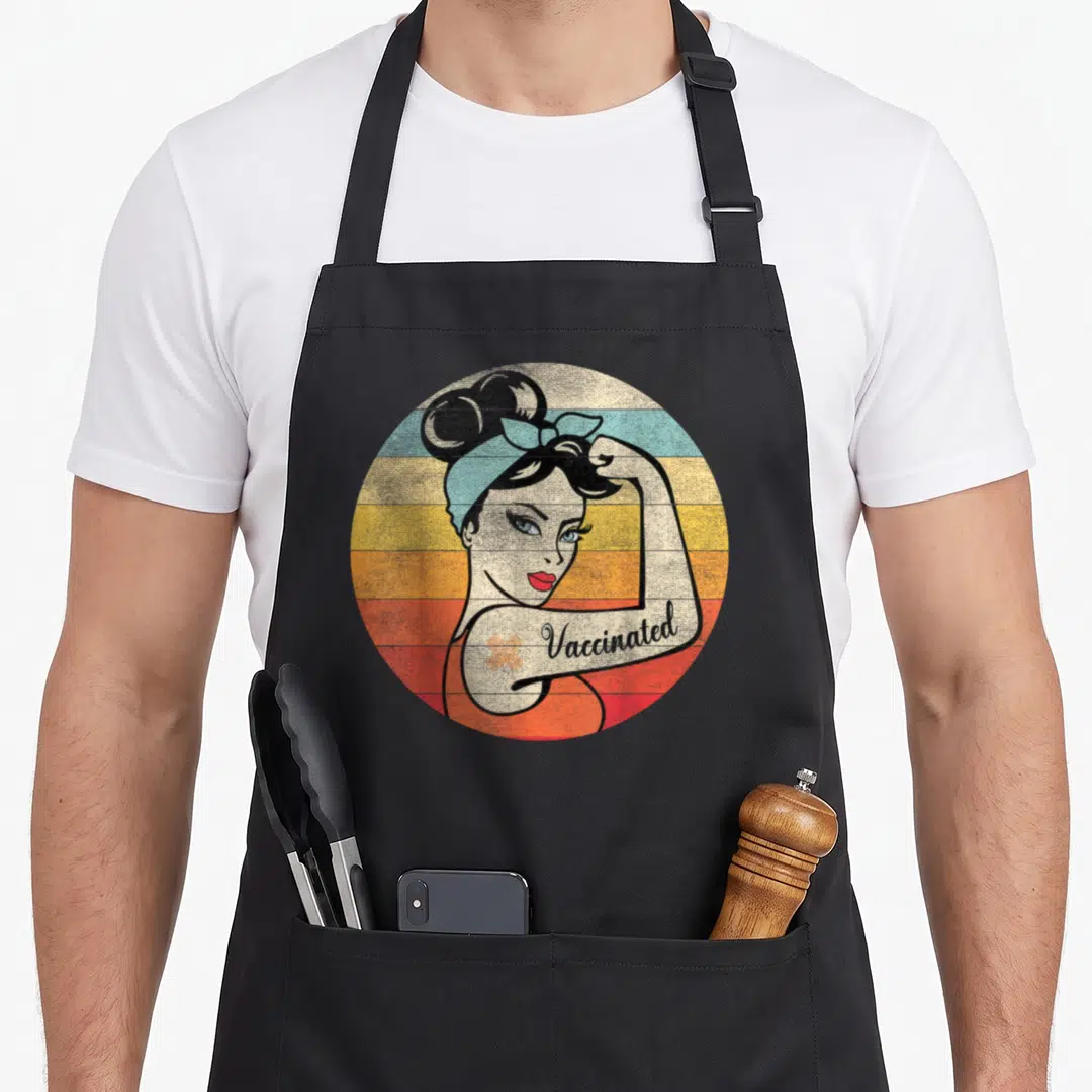 Believe vaccine apron - Retro woman flexing arm with Vaccinated text on colored stripes — shown on a black cotton canvas apron against a white background