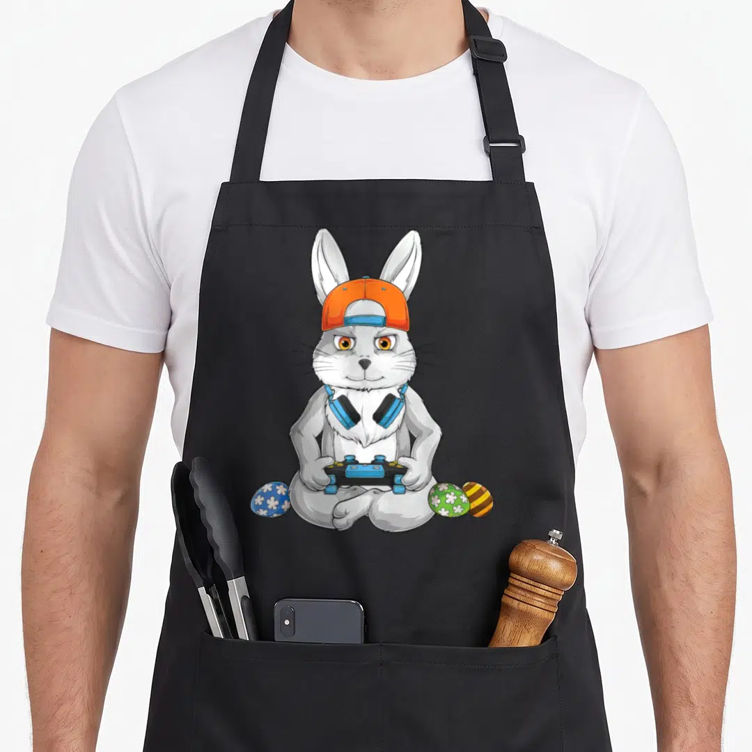Egg easter bunny aprons - White rabbit with orange cap, blue headphones, game controller, and three decorated eggs — shown on a black cotton canvas apron against a white background