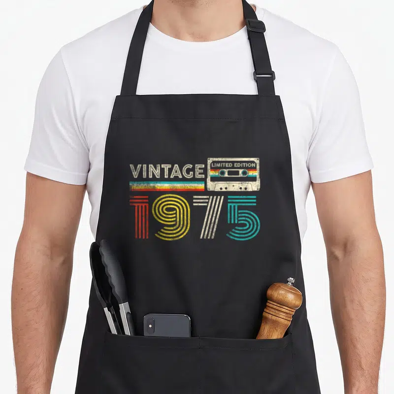 Funny cassette birthday apron - Retro cassette tape graphic with vintage 1975 text and colored stripes — shown on a black cotton canvas apron against a white background