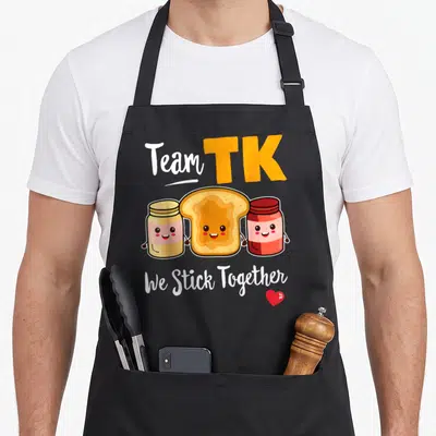 Transitional kindergarten teacher apron - Cartoon toast holding hands with peanut butter and jam jars under Team TK text — shown on a black cotton canvas apron against a white background
