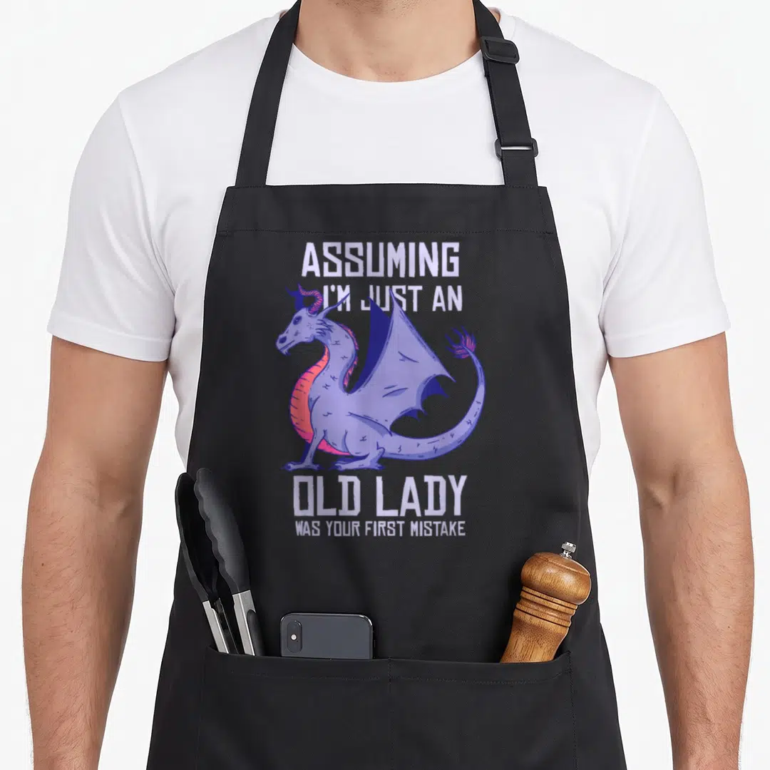 Dragon grandma apron - Purple cartoon dragon between white text reading Assuming I'm Just an Old Lady Was Your First — shown on a black cotton canvas apron against a white background