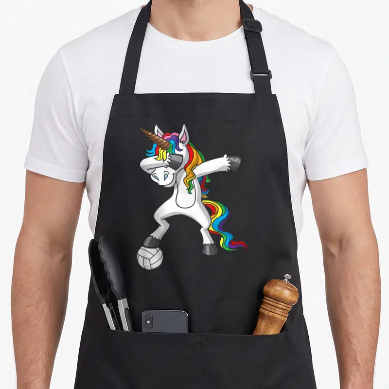Volleyball unicorn apron - White cartoon unicorn dabbing with rainbow mane and grey hooves standing on a volleyball — shown on a black cotton canvas apron against a white background