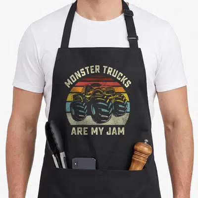 Retro monster truck apron - Retro monster truck centered on striped beige, red, orange, yellow, and blue sun with text — shown on a black cotton canvas apron against a white background