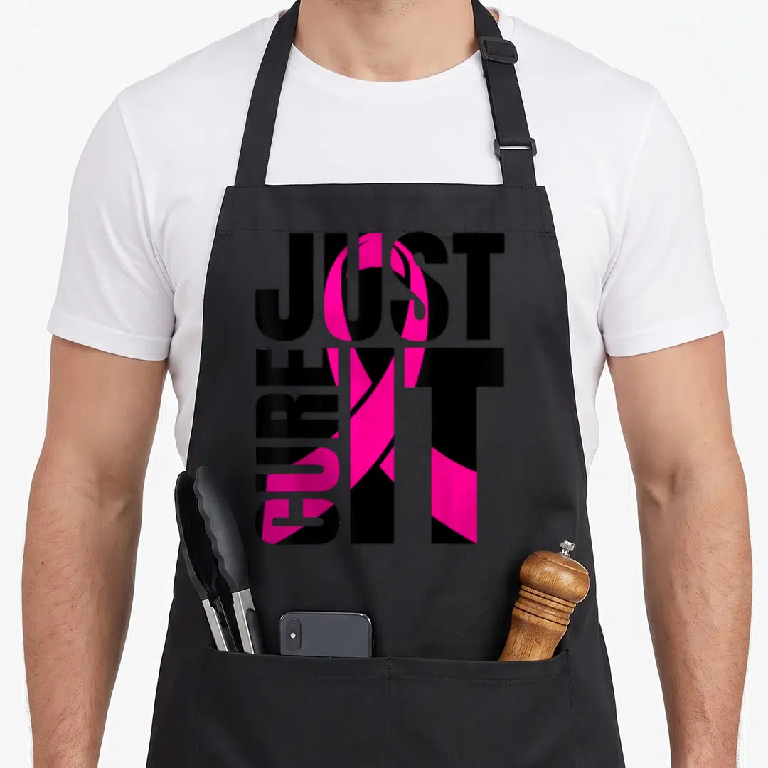 Cure breast cancer awareness aprons - Just Cure It text with pink ribbon overlapping black typography — shown on a black cotton canvas apron against a white background