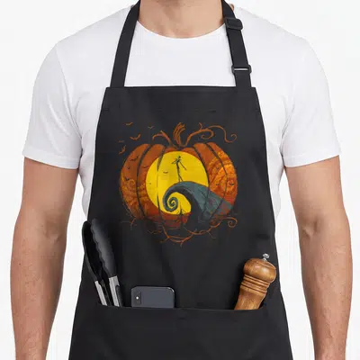 Lament pumpkin apron - Orange pumpkin frames yellow moon, black skeleton stands on grey swirl hill, scattered bats — shown on a black cotton canvas apron against a white background