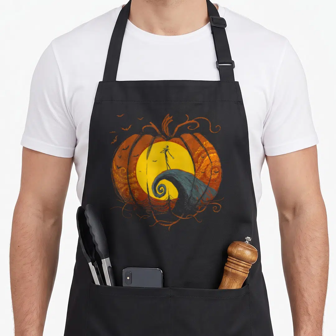 Lament pumpkin apron - Orange pumpkin frames yellow moon, black skeleton stands on grey swirl hill, scattered bats — shown on a black cotton canvas apron against a white background