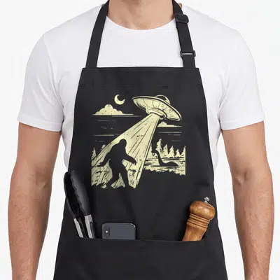Lochness bigfoot apron - Beige retro ufo beam lifting bigfoot silhouette near loch ness monster in forest with moon — shown on a black cotton canvas apron against a white background