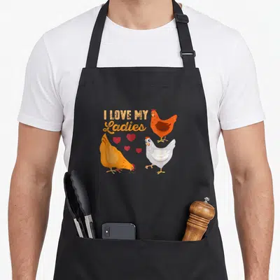 Funny farmer animal chicken apron - Three cartoon hens with four red hearts below I Love My Ladies text — shown on a black cotton canvas apron against a white background