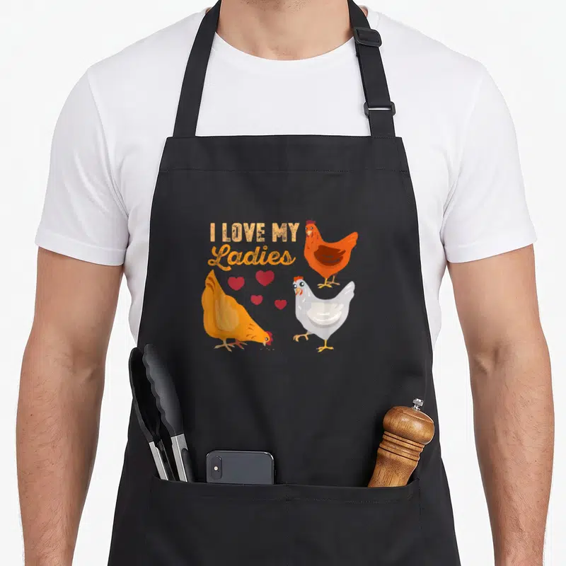 Funny farmer animal chicken apron - Three cartoon hens with four red hearts below I Love My Ladies text — shown on a black cotton canvas apron against a white background