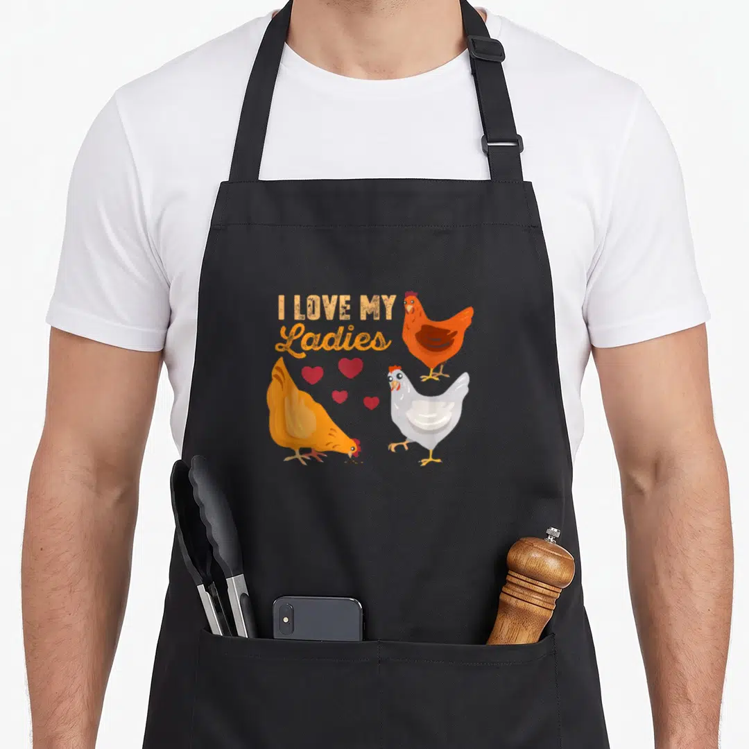 Funny farmer animal chicken apron - Three cartoon hens with four red hearts below I Love My Ladies text — shown on a black cotton canvas apron against a white background