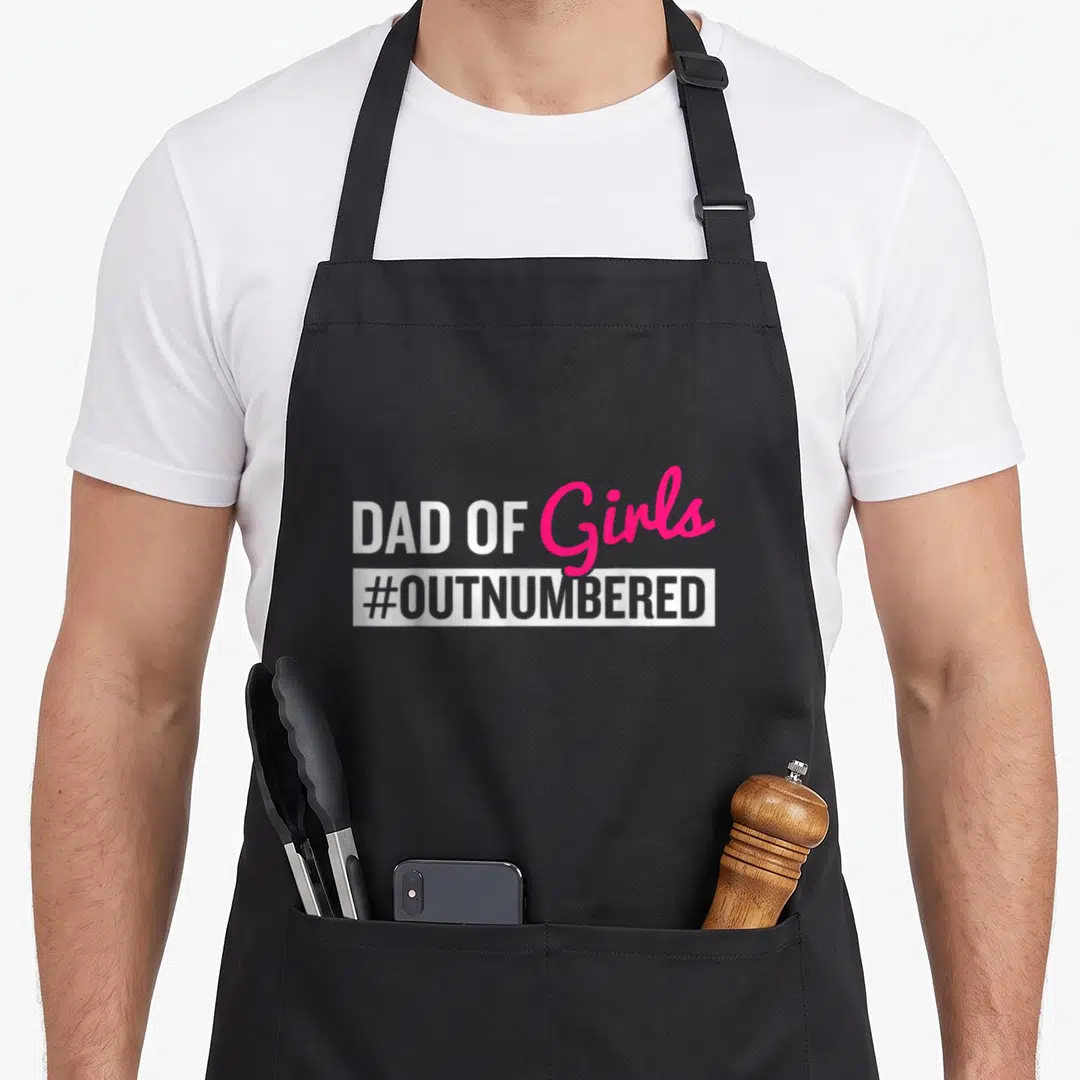 Outnumbered father day apron - DAD OF Girls #OUTNUMBERED text in white and pink typography on black fabric — shown on a black cotton canvas apron against a white background