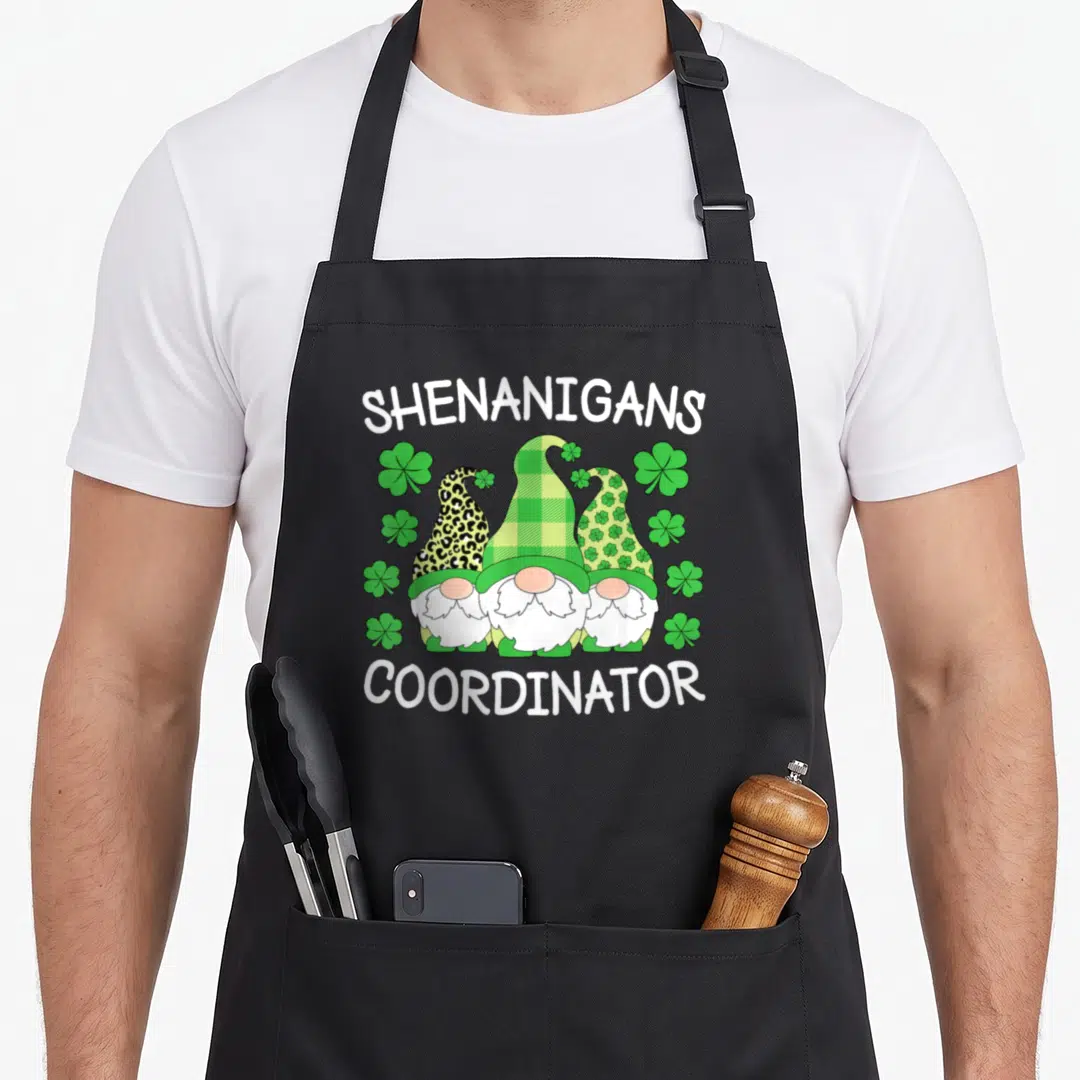 Teacher st patrick day aprons - Three gnomes with patterned hats surrounded by shamrocks and shenanigans coordinator — shown on a black cotton canvas apron against a white background