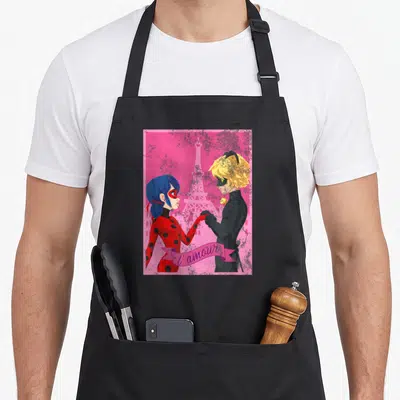 Miraculous paris apron - Two cartoon characters holding hands before Eiffel Tower with pink ribbon reading l'amour — shown on a black cotton canvas apron against a white background