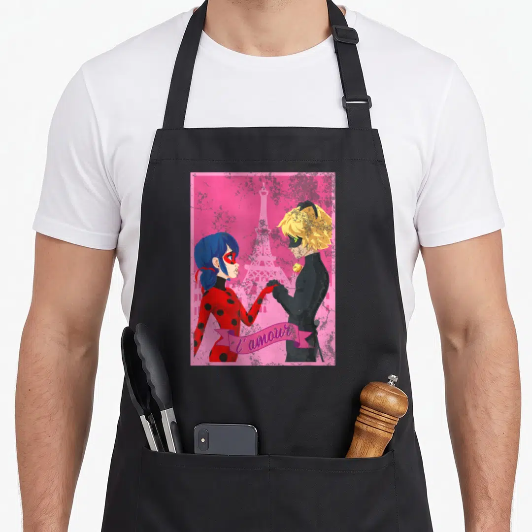 Miraculous paris apron - Two cartoon characters holding hands before Eiffel Tower with pink ribbon reading l'amour — shown on a black cotton canvas apron against a white background