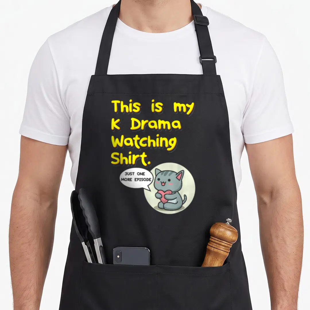 Drama watching apron - Cartoon grey cat holding red heart under yellow text reading K Drama Watching Shirt — shown on a black cotton canvas apron against a white background