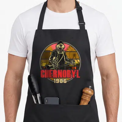 Chernobyl radiation aprons - Distressed hazmat figure with Geiger counter and Chernobyl 1986 text over ferris wheel — shown on a black cotton canvas apron against a white background