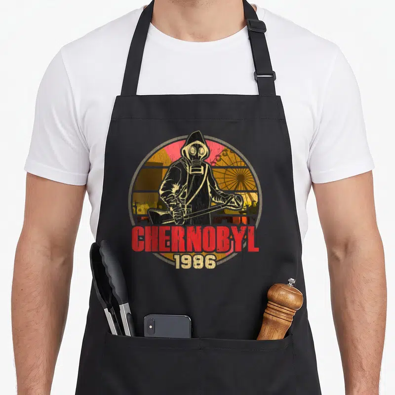 Chernobyl radiation aprons - Distressed hazmat figure with Geiger counter and Chernobyl 1986 text over ferris wheel — shown on a black cotton canvas apron against a white background