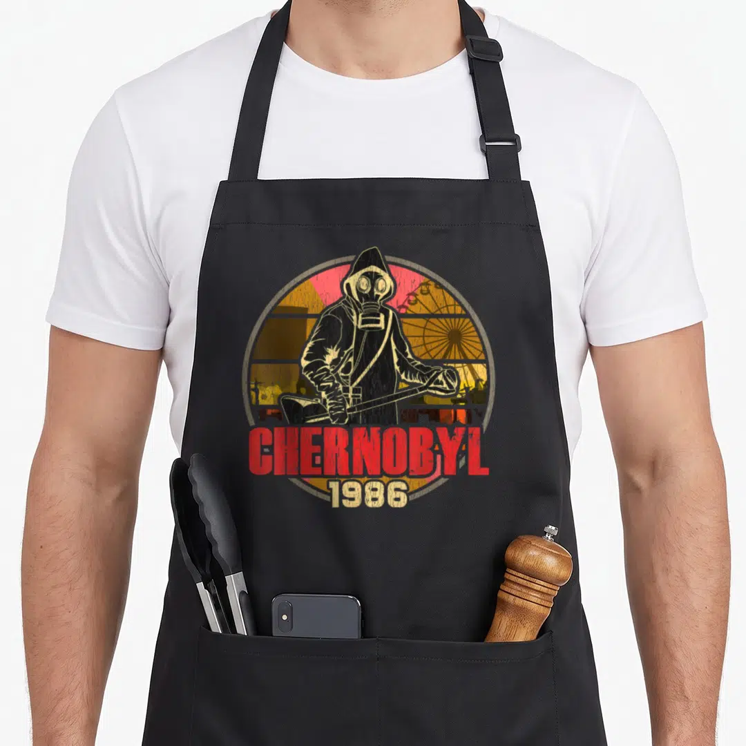 Chernobyl radiation aprons - Distressed hazmat figure with Geiger counter and Chernobyl 1986 text over ferris wheel — shown on a black cotton canvas apron against a white background