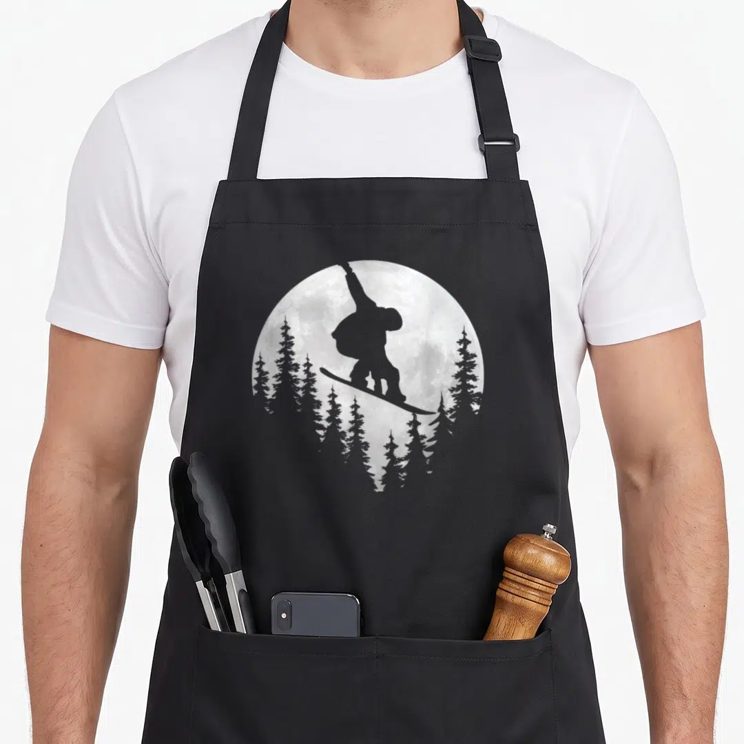 Snowboarding ski apron - Grey snowboarder silhouette jumping in front of a moon above pine trees — shown on a black cotton canvas apron against a white background