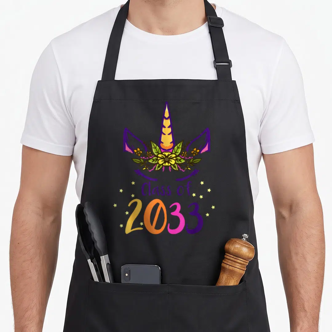 Face unicorn apron - Cartoon unicorn head with floral crown and Class of 2033 text surrounded by stars — shown on a black cotton canvas apron against a white background