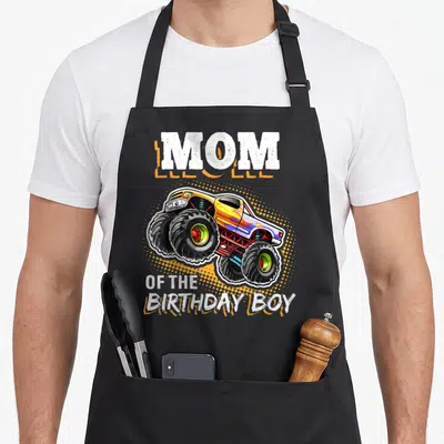 Vintage birthday monster truck apron - MOM OF THE BIRTHDAY BOY text with cartoon monster truck over orange halftone — shown on a black cotton canvas apron against a white background