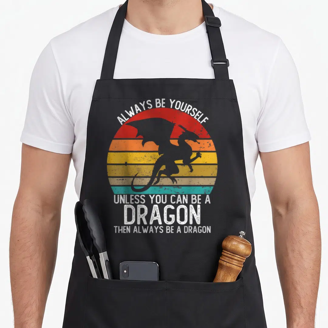 Unless dragon apron - Retro striped sun with grey dragon silhouette and white text reading Always Be Yourself Unless — shown on a black cotton canvas apron against a white background