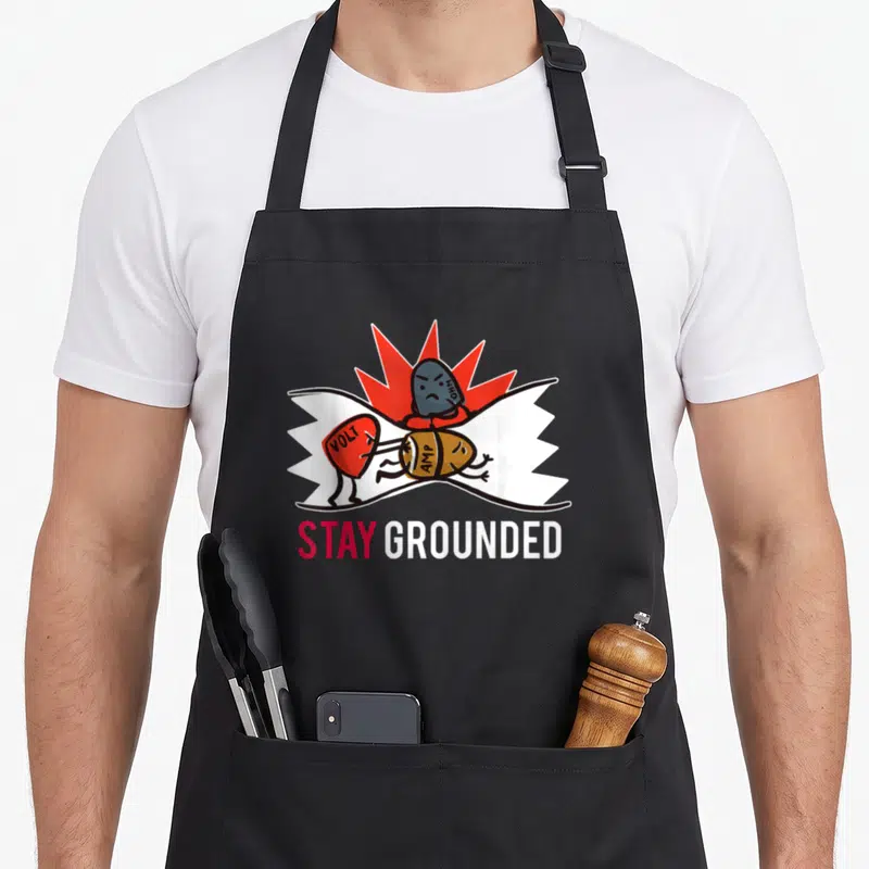 Master teacher apron - Cartoon VOLT, AMP, and OHM components on red spark background with STAY GROUNDED text — shown on a black cotton canvas apron against a white background
