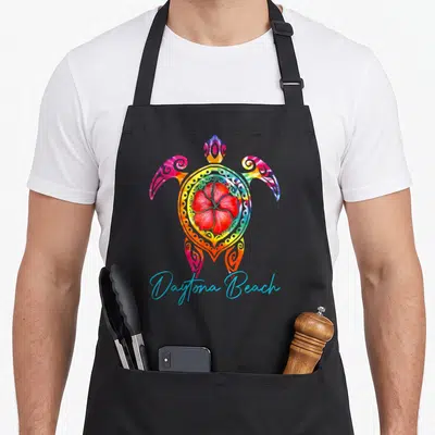 Daytona tie-dye apron - Multicolored sea turtle with red hibiscus shell and blue script text — shown on a black cotton canvas apron against a white background