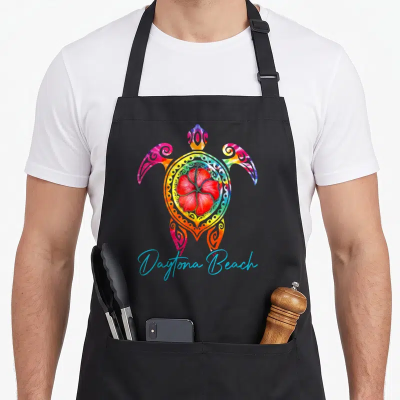 Daytona tie-dye apron - Multicolored sea turtle with red hibiscus shell and blue script text — shown on a black cotton canvas apron against a white background