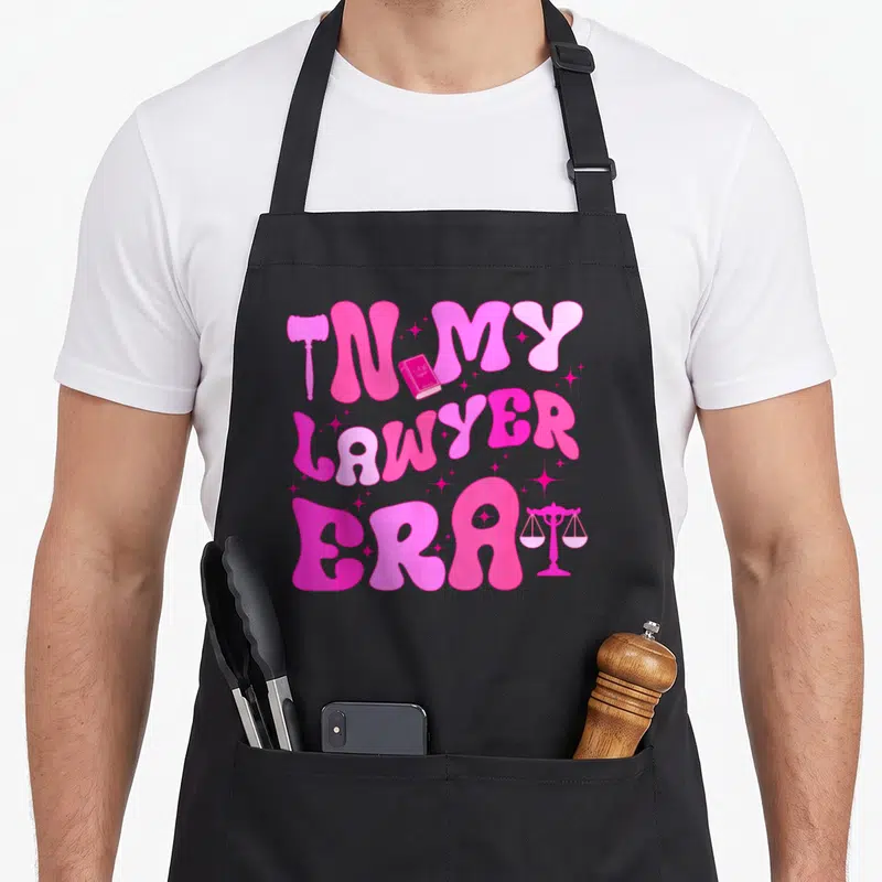 Groovy lawyer apron - Pink bubbly text IN MY LAWYER ERA with gavel book and scales — shown on a black cotton canvas apron against a white background