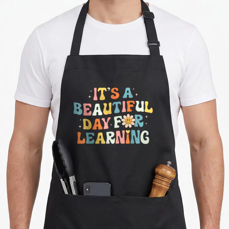 Funny beautiful teacher apron - Stacked retro text IT'S A BEAUTIFUL DAY FOR LEARNING with smiley flower and scattered — shown on a black cotton canvas apron against a white background