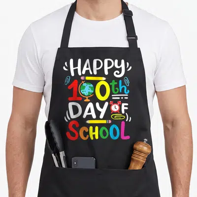 Student teacher apron - Happy 100th Day of School text with globe, pencils, and alarm clock in cartoon style — shown on a black cotton canvas apron against a white background