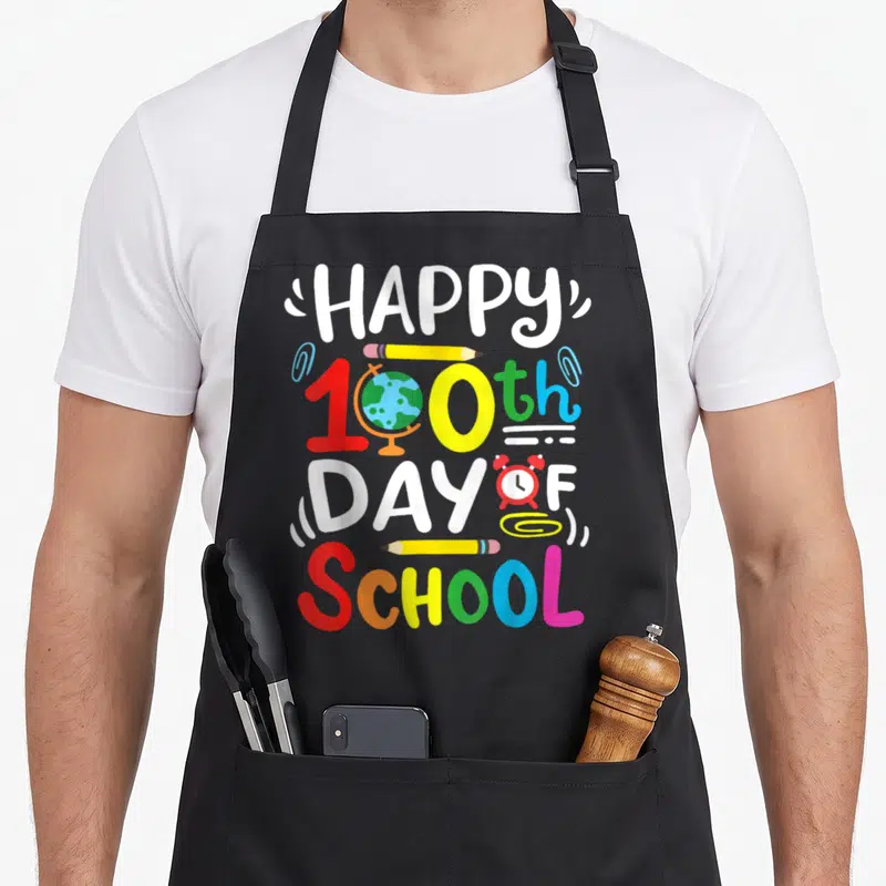 Student teacher apron - Happy 100th Day of School text with globe, pencils, and alarm clock in cartoon style — shown on a black cotton canvas apron against a white background