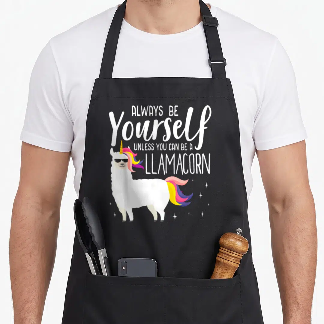 Alway magic apron - Cartoon llamacorn with sunglasses and rainbow mane under stacked white text about being a llamacorn — shown on a black cotton canvas apron against a white background